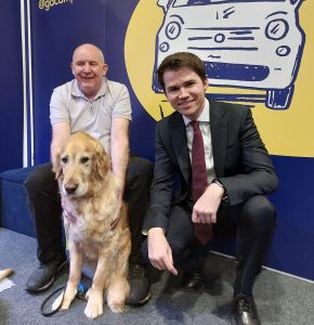 Paul is sat on a bench with Pyrgo in front of him. Gordon McKee, MP for Glasgow South, is kneeling to the right of Paul. The Guide Dogs “Pave the way for clearer streets” message is behind them on an illustrated backdrop.