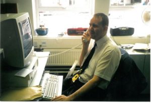Image of Paul sat on the phone in front of a computer wearing a smart shirt and black Disney character tie