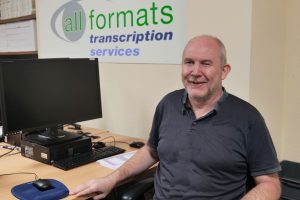 Image of Paul in 2026 wearing a grey polo smiling at his desk