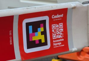Close up image of a red sticker which has a logo, QR code and writing for accessible signage